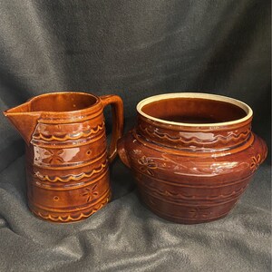 Puede incluir: Un juego de dos piezas de cocina de cerámica esmaltada marrón. Una jarra con asa y una olla redonda con tapa.