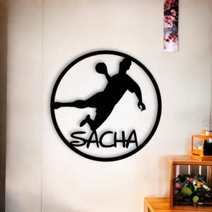 Peut inclure: Décoration murale en métal noir représentant un joueur de handball en plein saut, tenant le ballon. Le joueur est à l'intérieur d'un cercle noir, avec le nom "SACHA" en dessous. L'art est fixé sur un mur blanc.