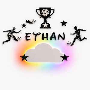 Peut inclure: Décoration murale sur le thème du football. Elle comprend un trophée, des ballons de foot, des étoiles et des silhouettes de joueurs. Le nom "ETHAN" est affiché au-dessus d'un nuage lumineux avec un effet arc-en-ciel.