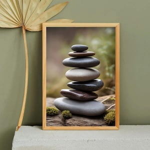 May include: A framed print of a stack of smooth, gray and brown stones balanced on top of each other. The stones are arranged in a pyramid shape and are set against a blurred background of green foliage.