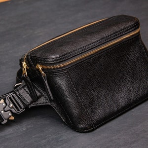 以下が含まれることがあります： 黒い革製のウエストバッグで、金色のジッパーと黒いバックルが付いています。バッグは濃い灰色の表面の上にあります。