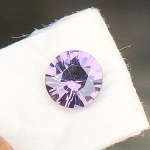 May include: A round, faceted gemstone with a light purple hue. The gem is set on a white cloth, showcasing its brilliant cut and reflective surfaces. The gemstone's facets create a starburst effect, highlighting its clarity and color.