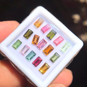 May include: A white tray with 15 small, rectangular, faceted gemstones in various colors, including green, pink, red, orange, and yellow.