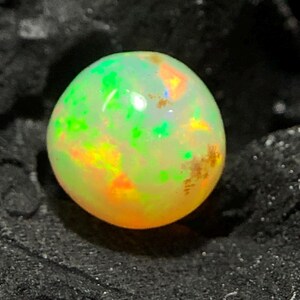 May include: A round opal gemstone with a vibrant display of colors. The opal features a mix of green, yellow, and orange hues, creating a luminous effect. The gemstone is set against a dark, textured background.