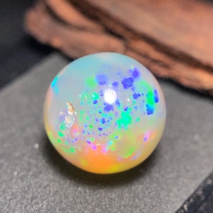 May include: A round opal gemstone with a white base, displaying vibrant flashes of blue, green, and orange. The gemstone is polished and smooth, with a subtle iridescence. The opal is set against a dark background.