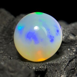 May include: A round, iridescent opal gemstone with a milky white base color. The opal displays flashes of blue, green, and orange hues. The gemstone is set against a dark, textured background.