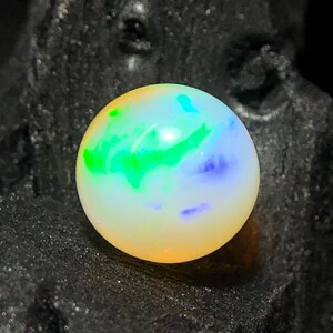 May include: A round, iridescent gemstone with a mix of colors, including green, blue, and yellow. The gemstone is set against a dark, textured background, highlighting its vibrant hues and spherical shape. The gem appears to be an opal.
