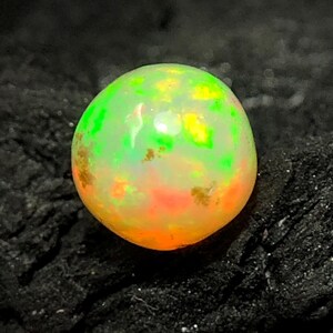 May include: A round opal gemstone with a vibrant display of colors. The opal features a mix of green, yellow, and orange hues, creating a luminous effect. The gemstone is set against a dark, textured background.