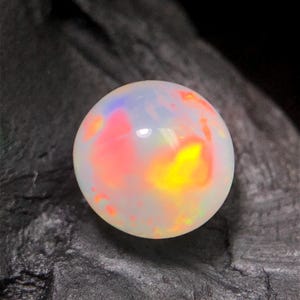 May include: A round, polished opal gemstone featuring a translucent white base with vibrant flashes of red, orange, and yellow play-of-color. The surface of the stone is smooth and reflective, showcasing internal light refractions throughout.
