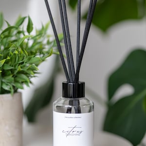 May include: A clear glass reed diffuser with black reeds. The diffuser has a black cap and a white label with the words "Citrus Sunshine". The diffuser is 118 ml and is next to a small green plant.