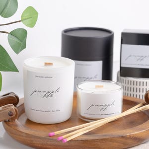 Puede incluir: Velas blancas con tapas de madera y el texto "pineapple sage" se exhiben en una bandeja de madera. Las velas están en recipientes de vidrio y cerámica. Fósforos con puntas rosas están junto a las velas. Una caja cilíndrica negra está en el fondo.