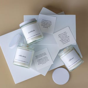 Op de afbeelding: Vier glazen potkaarsen met witte etiketten met de tekst "dankbaarheid", "reflectie" en "zelfliefde". De kaarsen staan op witte geometrische vormen met inspirerende citaten. De deksels zijn zilver en wit.