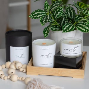 Puede incluir: Una exhibición de velas perfumadas y objetos decorativos. La imagen muestra un recipiente negro para velas, dos velas blancas en vasos y una bandeja de madera. Las velas tienen la inscripción "sea salt orchid". Una guirnalda de cuentas y una planta en maceta completan la escena.