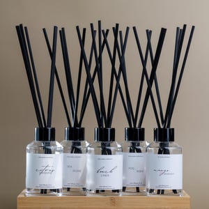 Peut inclure: Ensemble de cinq diffuseurs à roseaux en verre transparent avec des roseaux noirs, chacun étiqueté avec un parfum différent : Citrus Sunshine, Sea Stone, Beach Linen, Fog Fern et Mango Grapefruit. Les diffuseurs sont présentés dans un plateau en bois.