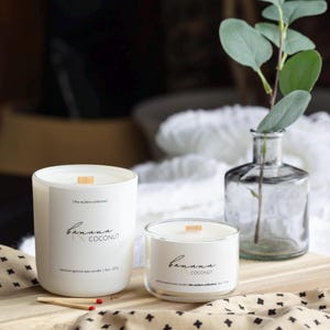 May include: Two white glass candles with wooden wicks, labelled "Banana Coconut." The larger candle is 227g, the smaller is 113g. A clear glass vase with green leaves and matches are on a wooden surface.
