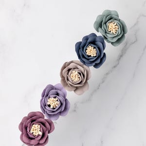 May include: Five fabric flowers are arranged diagonally, viewed from above. The flowers are in shades of purple, mauve, taupe, navy, and teal. Each flower has a textured, cream-coloured centre. The background features a white marble pattern.