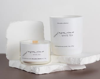 May include: Two white coconut apricot wax candles with wooden wicks. The smaller candle is labelled "jasmine white tea (the auders collective) 4oz - 113g". The larger candle is labelled "jasmine white tea (the auders collective) 8oz - 227g".