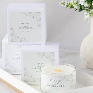 Op de afbeelding: Witte kaars in een doorzichtige glazen container met een houten lont, op een wit dienblad. De kaars en dozen zijn voorzien van de namen "Olivia en Alexander" en de datum "07.17.27", met bloemmotieven. Huwelijksbedankjes.