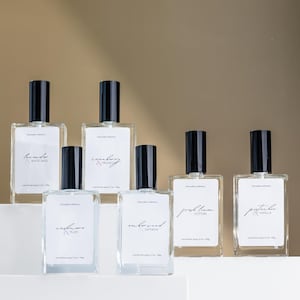 May include: Six clear glass bottles with black spray tops and white labels. The labels have handwritten text describing the scents: "Honeysuckle & White Sage", "Cranberry & Prosecco", "Cashmere & Plum", "Amberwood & Saffron", "Fresh Linen & Cotton", and "Pistachio & Vanilla".