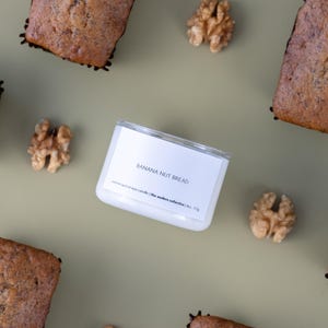 May include: A white candle labelled "BANANA NUT BREAD" is surrounded by slices of banana nut bread and walnuts on a green surface. The candle is labelled "coconut apricot wax candle | the auders collective | 113g".