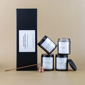 Op de afbeelding: Een zwarte rechthoekige doos met een wit etiket met de tekst "THE SEASONAL CANDLE COLLECTION". Vier glazen potten met zwarte deksels en etiketten zijn gestapeld. Een koperen kaarsenstopper en een lange koperen staaf zijn ook aanwezig.