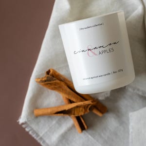Puede incluir: Una vela blanca con las palabras "cinnamon & apples" en letra cursiva. La vela está sobre una tela gris claro junto a varios palitos de canela. La vela es de 227g.