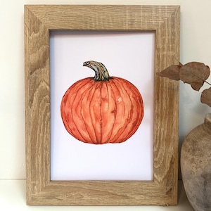May include: Watercolour painting of a round orange pumpkin with a green and brown stem, set in a light brown wooden frame. The artwork is on a white background, evoking an autumn or harvest theme.