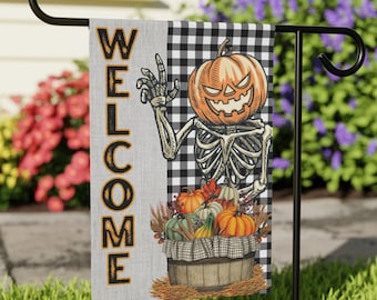 Op de afbeelding: Een Halloween-thema welkomsvlag met een skelet dat een pompoen vasthoudt en een mand met pompoenen. De vlag is zwart-wit geruit met het woord "WELCOME" in zwarte letters.