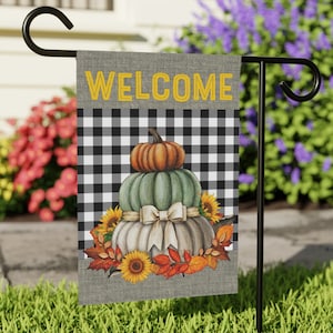 May include: A welcome garden flag with a black and white checked background. The flag features a stack of three pumpkins with a white ribbon and autumn leaves. The top pumpkin is orange, the middle pumpkin is green, and the bottom pumpkin is white.
