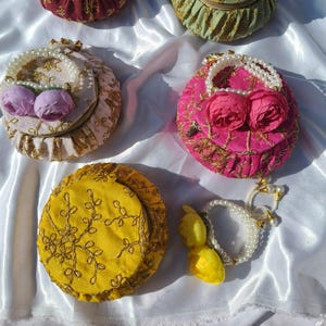Peut inclure: Plusieurs boîtes rondes décoratives en tissu, de couleurs variées : bordeaux, vert, rose et jaune. Chaque boîte est ornée de motifs floraux et d'une poignée perlée. Les boîtes sont disposées sur un tissu blanc satiné.