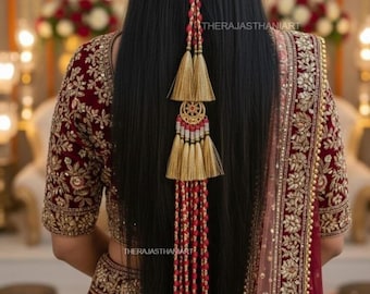 May include: A woman's back is shown, wearing a maroon and gold embroidered outfit. A long hair accessory with gold tassels and red beaded accents is attached to the hair. The text "THERAJASTHANIART" is visible.