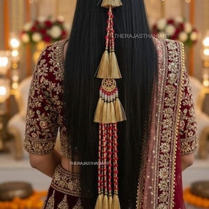 Puede incluir: Vista trasera de una mujer con un atuendo bordado en burdeos y dorado. Un largo accesorio para el cabello con borlas doradas y detalles de cuentas rojas está sujeto al cabello. El texto "THERAJASTHANIART" es visible.
