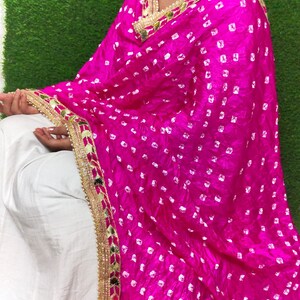 May include: A pink silk dupatta with white polka dots and a gold and pearl border. The dupatta is draped over a white outfit.