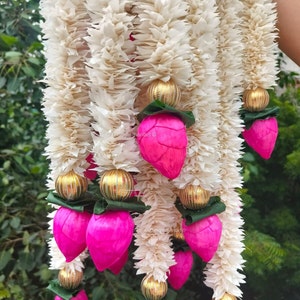 Pode incluir: Uma grinalda decorativa com flores de papel texturizado branco sujo. Acentuada com ornamentos esféricos dourados e elementos rosa vibrantes em forma de lótus com folhas verdes. A grinalda é exibida contra um fundo de folhagem verde.
