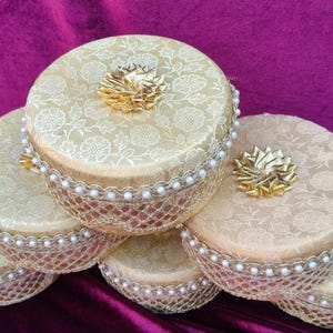 Puede incluir: Una pila de cajas redondas y decorativas con tapas estampadas con flores. Las cajas son de color beige con detalles dorados, incluyendo un lazo dorado en la parte superior y un ribete con cuentas de perlas. Las cajas están apiladas sobre una superficie de terciopelo morado oscuro.