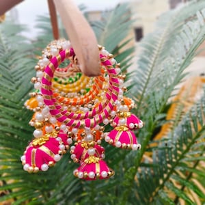 以下が含まれることがあります： ピンク、オレンジ、イエロー、グリーンの生地と白いパールをあしらったカラフルなバングルセット。バングルは小さなピンクのファブリックタッセルで飾られています。