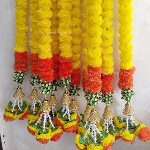 Puede incluir: Una guirnalda vibrante con flores amarillas y naranjas, con detalles verdes y dorados. La guirnalda está adornada con campanas decorativas y es adecuada para ocasiones festivas. El texto "Therajasthaniart" es visible.