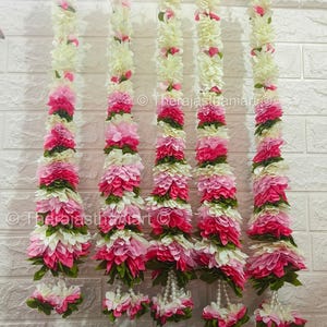 Puede incluir: Cinco guirnaldas de flores rosas y blancas con hojas verdes y detalles de perlas colgando de una pared de ladrillo blanco.