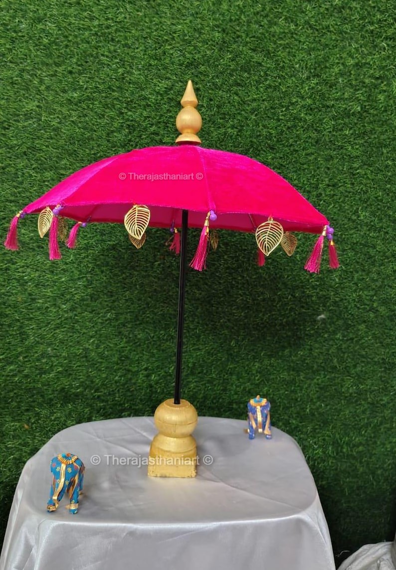 K&ouml;nnte beinhalten: Ein rosa Samt-Schirm mit goldenen Blatt-Akzenten und rosa Quasten. Der Schirm steht auf einem Holzst&auml;nder mit goldener Basis.