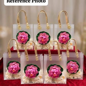 Könnte beinhalten: Sieben transparente Tragetaschen mit goldenen Griffen, jeweils mit einer rosa Lotusblume und weißen floralen Akzenten verziert. Die Taschen sind auf einer roten Oberfläche angeordnet. Der Text "Reference Photo" befindet sich oben.