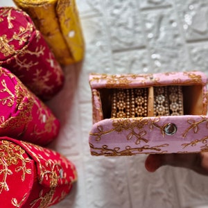 Puede incluir: Caja de joyería bordada en rosa y dorado con una tapa con bisagras abierta, revelando dos filas de brazaletes de oro y plata en el interior.