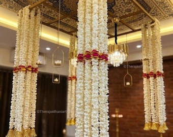 May include: A ceiling-mounted decorative installation featuring multiple hanging strands of white jasmine flowers accented with magenta floral bands and gold-toned bell-shaped ornaments at the base. The structure includes a rectangular frame.
