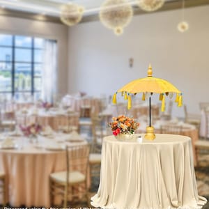 Può includere: Un ombrello di tessuto giallo con nappe blu si trova su un tavolo con una tovaglia bianca. Un piccolo mazzo di fiori è in un vaso bianco sul tavolo. L'ombrello è un accento decorativo per un matrimonio o una festa.