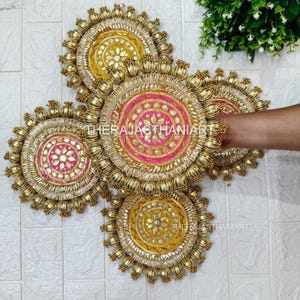 May include: Five decorative, circular wall hangings. Each features a central design in pink or yellow, surrounded by gold trim and embellishments. The text "THERAJASTHANIART" is visible on some of the pieces. The hangings are arranged against a white brick wall.