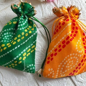 Può includere: Due sacchetti con coulisse e motivi bandhani. Uno è verde con dettagli gialli, l'altro è arancione con dettagli rossi. Entrambi hanno finiture color oro sulla parte superiore e cordini abbinati.