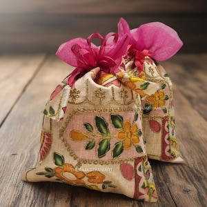 Op de afbeelding: Twee decoratieve stoffen zakjes met bloemenborduurwerk in roze, geel en groen. De zakjes zijn vastgebonden met roze linten en liggen op een houten oppervlak. De tekst "THERAJASTHANIART" is zichtbaar.