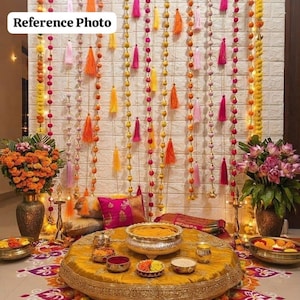 May include: A decorative display featuring a mustard yellow circular floor cushion with gold embroidery, surrounded by brass bowls, floral arrangements, and a backdrop of hanging strings of colorful pom-poms and tassels with the text Reference Photo.