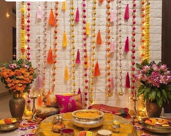 Könnte beinhalten: Ein dekoratives Arrangement mit einem senfgelben, runden Bodenkissen mit Goldstickerei, umgeben von Messingschalen, Blumenarrangements und einem Hintergrund aus hängenden Schnüren mit bunten Pompons und Quasten mit dem Text Reference Photo.
