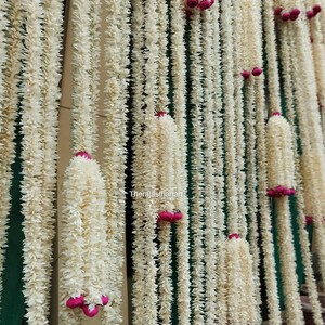 Op de afbeelding: Close-up van een decoratieve slinger gemaakt van wit, getextureerd materiaal, mogelijk bloemen of stof. De slinger is versierd met kleine, roze accenten. De tekst "therajasthanart" is zichtbaar. De slingers hangen verticaal.