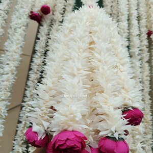 Op de afbeelding: Een close-up van een witte bloemenslinger met roze accenten. De slinger is gemaakt van gelaagde, getextureerde witte bloemblaadjes en heeft clusters van levendige roze bloemen. De slinger hangt verticaal.